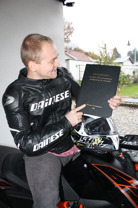 Johannes Stockhammer in a motorcycle jacket, sitting on a motorcycle and pointing at his bound diploma thesis