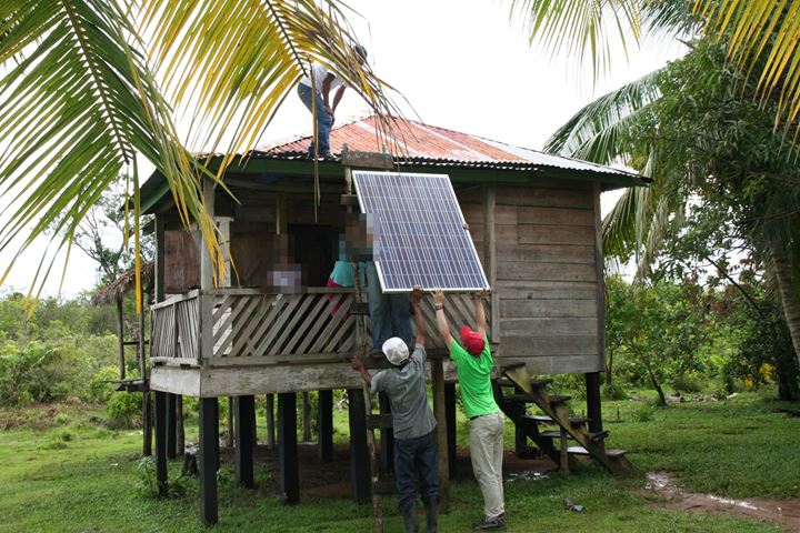 Team members lifting a solar panel onto a wooden house on stilts in Kukalaya